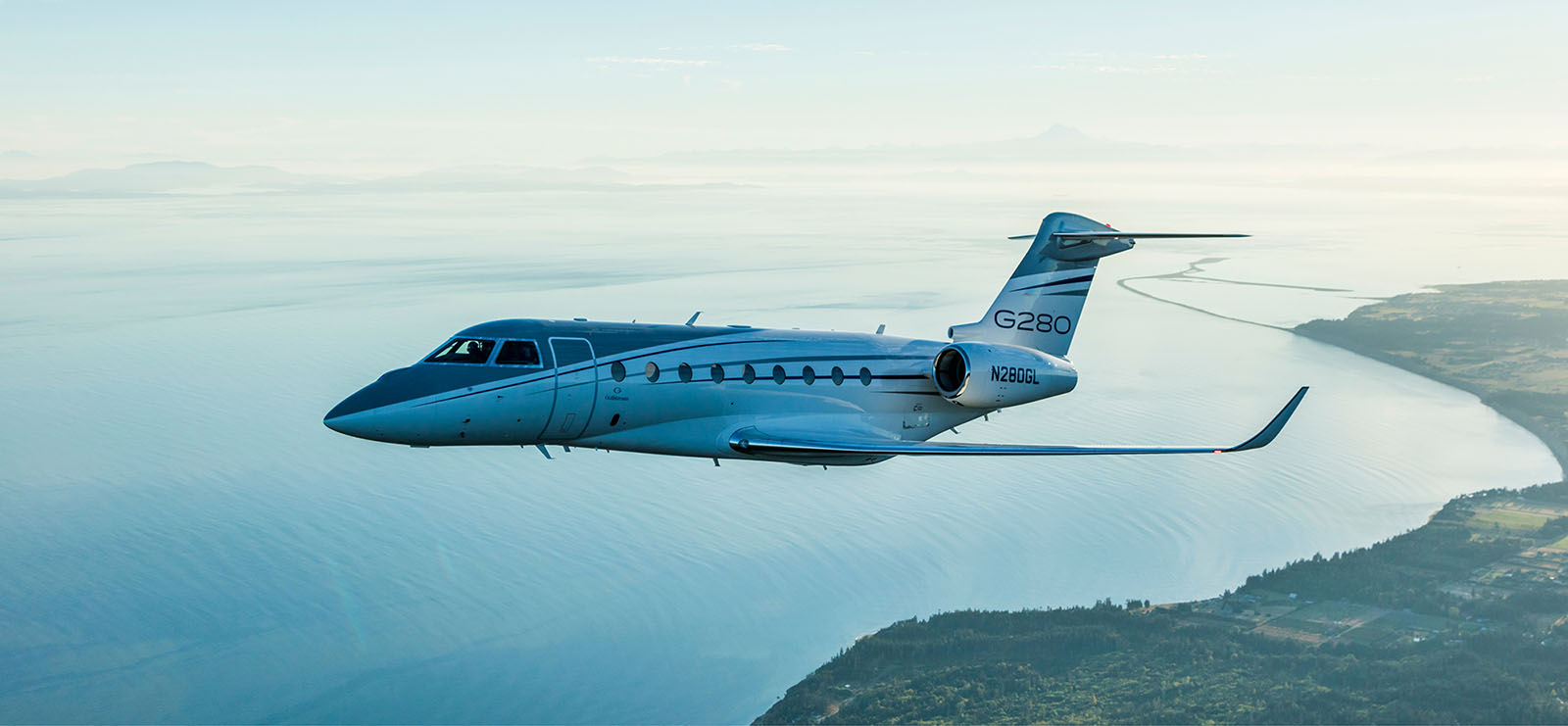 A Gulfstream G280 ascends over a green-tinted coastland and calm seas in morning sunlight
