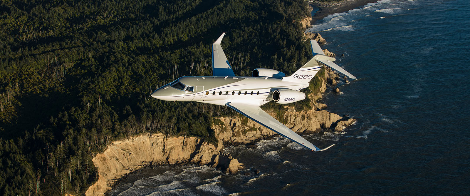 G280 ascending in bright morning sunlight over a rugged, forested coast