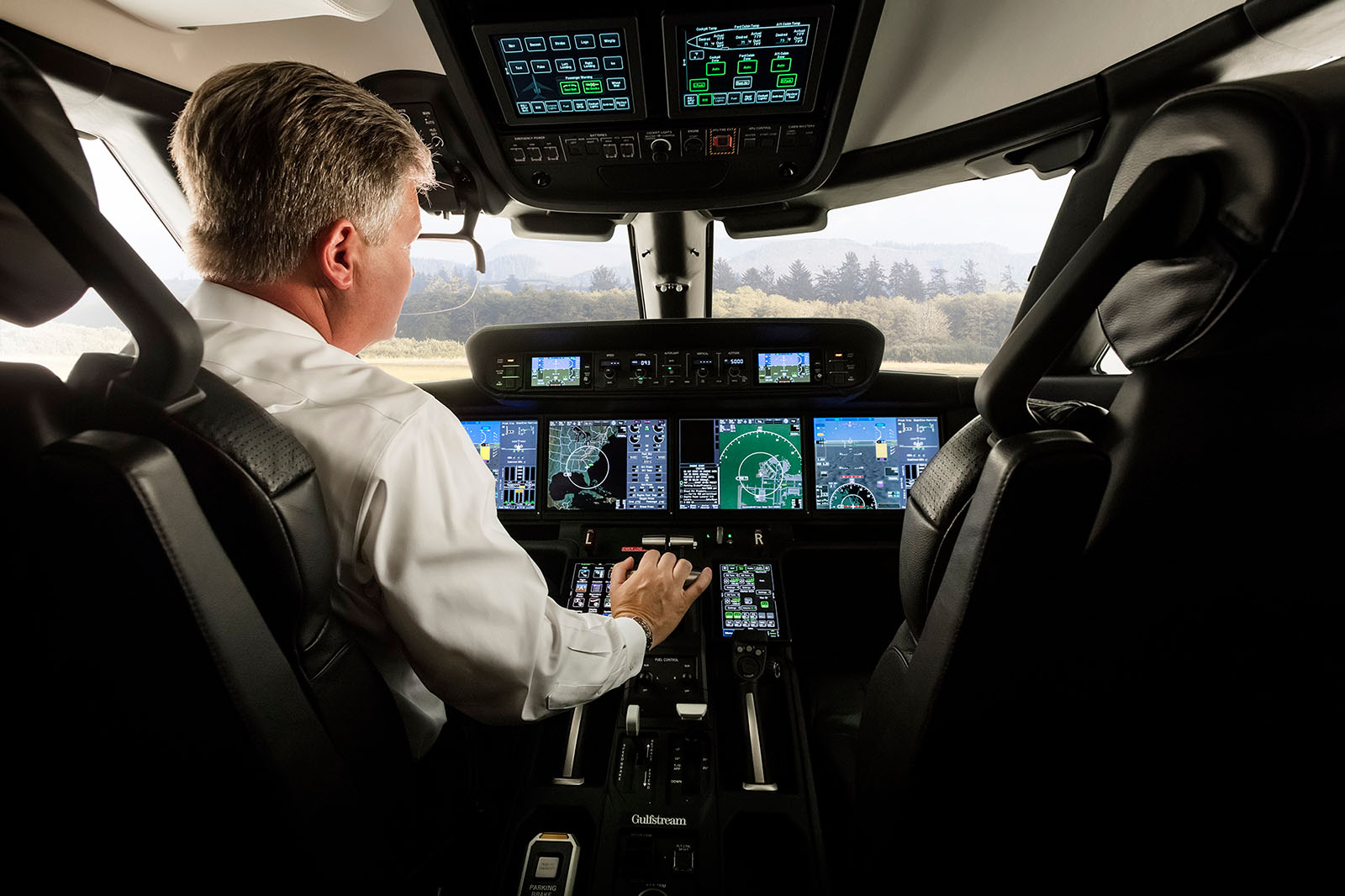 A view in the Gulfstream G400 flight deck showing a pilot with hands on the center throttle and a tree-lined airport out the window