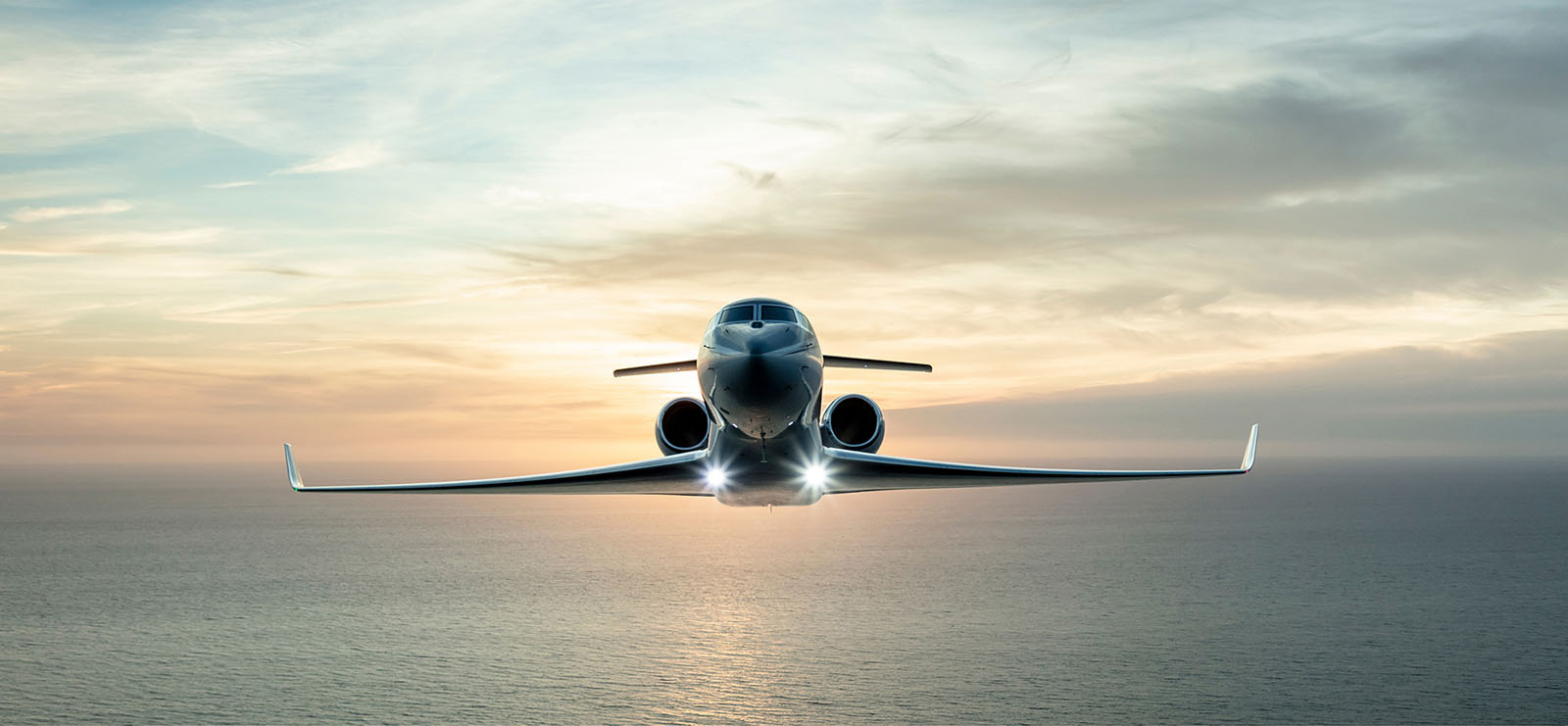 A backlit Gulfstream G600 flies directly toward the viewer over calm water at sunset