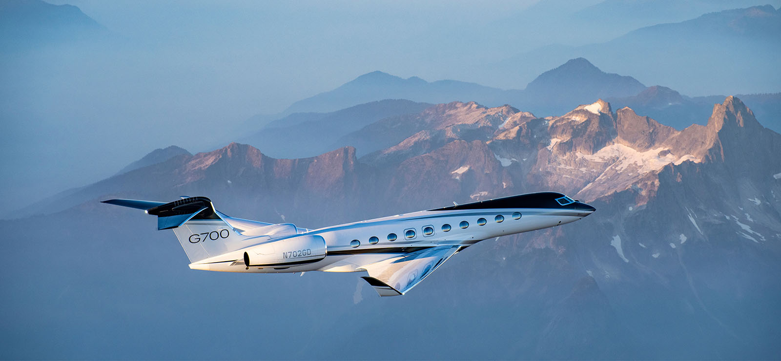 A Gulfstream G700 aircraft seen from slightly above and behind flies over rocky, snow-covered mountain peaks.