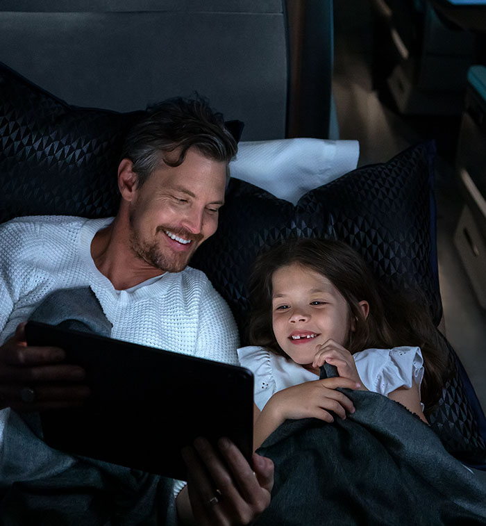 A father and daughter are bathed in the the light of a tablet in a darkened cabin