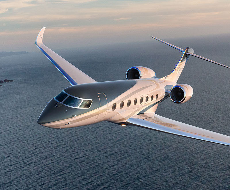 A Gulfstream G800 viewed from the front left quarter flies low over calm blue coastal waters.